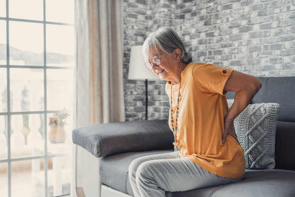 Unzufriedene reife Frau, die den Rücken berührt, Schmerzen fühlt, auf der Couch im Wohnzimmer sitzt, ungesunde Seniorin mittleren Alters, die unter Rückenschmerzen, Rückenproblemen, Reiben steifen Muskeln oder Nieren leidet - Foto, Bild