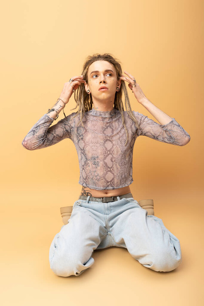 Queer person in crop top with snakeskin print and jeans touching dreadlocks on yellow background  - Photo, image