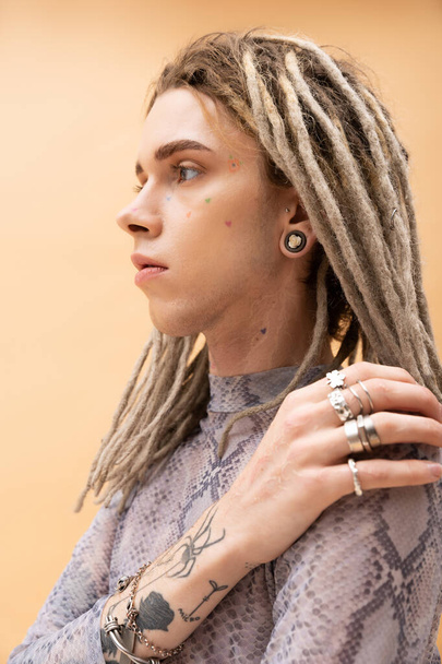 Portrait of tattooed queer person with dreadlocks touching shoulder isolated on yellow  - Fotografie, Obrázek