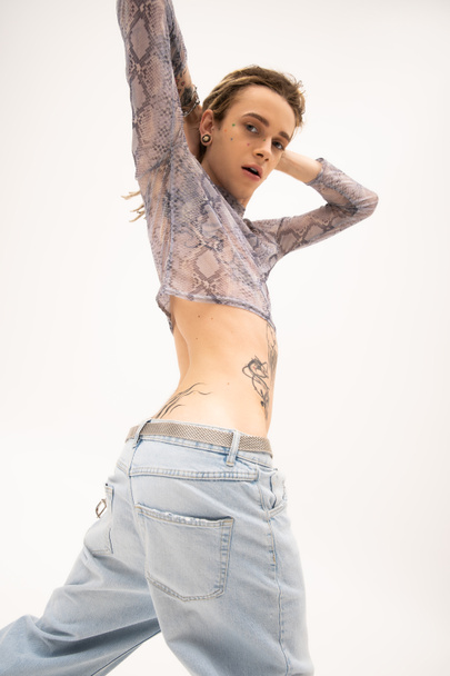 Low angle view of tattooed queer person in crop top with animal print looking at camera isolated on white  - Fotó, kép