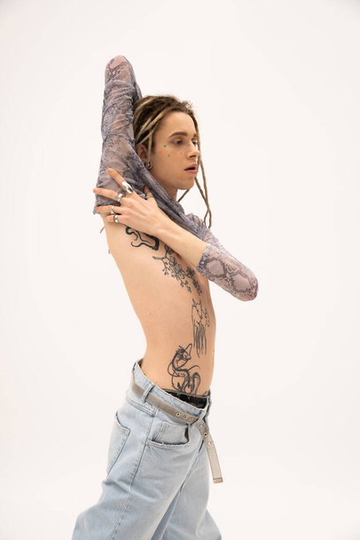 Tattooed queer person in jeans touching crop top isolated on white  - Fotó, kép