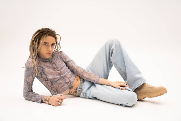 Tattooed nonbinary person in crop top with snakeskin print and jeans lying on white background - Photo, Image