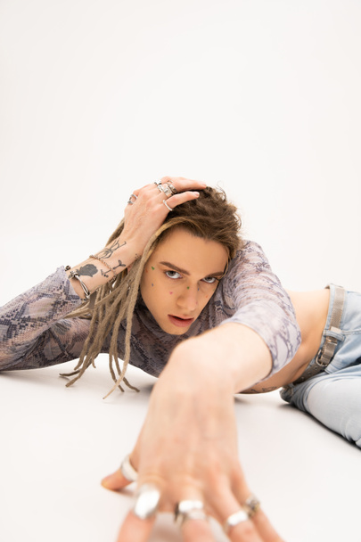 Tattooed nonbinary person looking at camera while lying on white background - Foto, Imagen