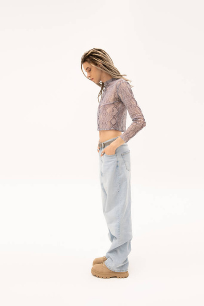 Side view of stylish queer person in crop top and jeans standing on white background - Foto, Imagen