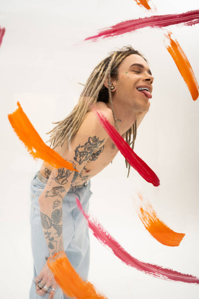 positive nonbinary person with tattooed body sticking out tongue near colorful paint spills on white background - Fotó, kép