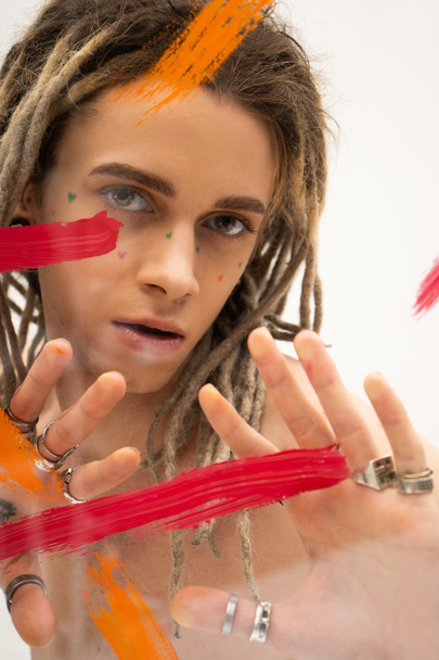 portrait of queer model with dreadlocks and silver rings looking at camera near paint strokes on white background - Foto, Bild
