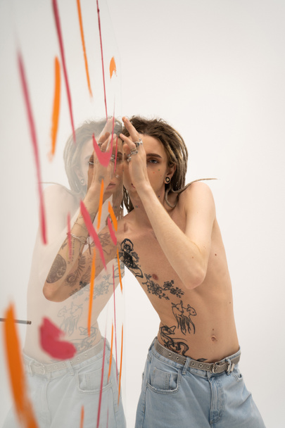 shirtless tattooed queer person in jeans posing near glass with colorful paint on white background - Photo, image