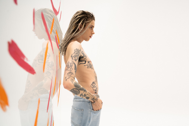 side view of shirtless tattooed nonbinary model posing with hand in pocket of jeans near glass with paint strokes on white background - Фото, изображение