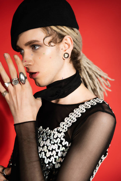 nonbinary model with dreadlocks and silver finger rings holding hands near face and looking away on red  - Zdjęcie, obraz