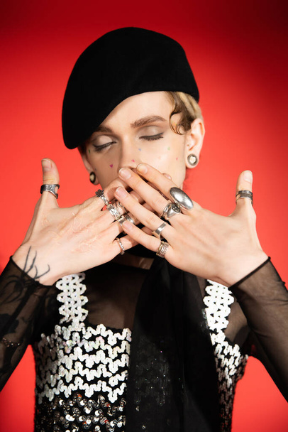 queer person with closed eyes covering mouth with hands in silver rings on red background - Foto, Bild