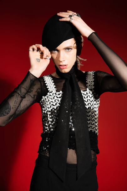 tattooed queer person in top with sequins adjusting black beret and looking at camera on red background - Zdjęcie, obraz