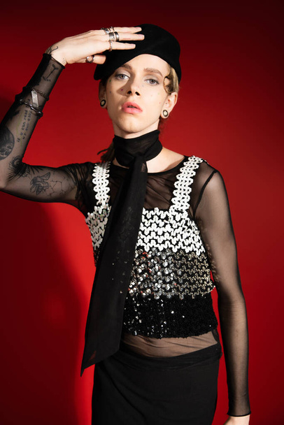 tattooed queer person in elegant top and silver rings adjusting black beret on red background - Photo, Image