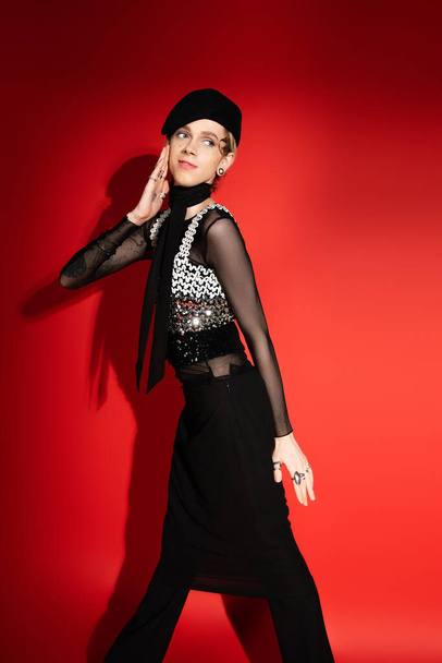 young queer model in elegant outfit posing with hand near face on red background with shadow - Fotó, kép