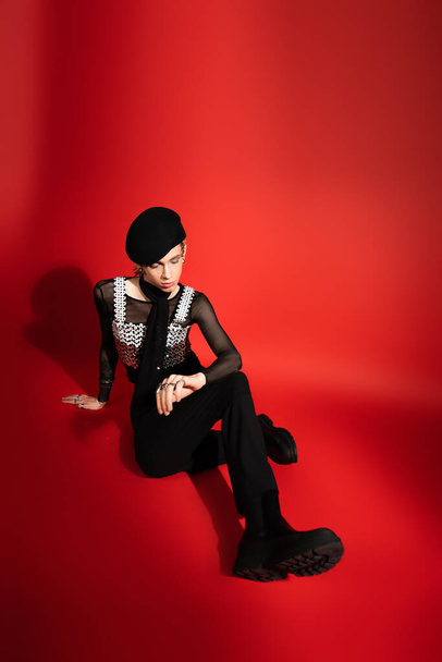 full length of young nonbinary person in black beret and top with sequins sitting on red background - Fotoğraf, Görsel