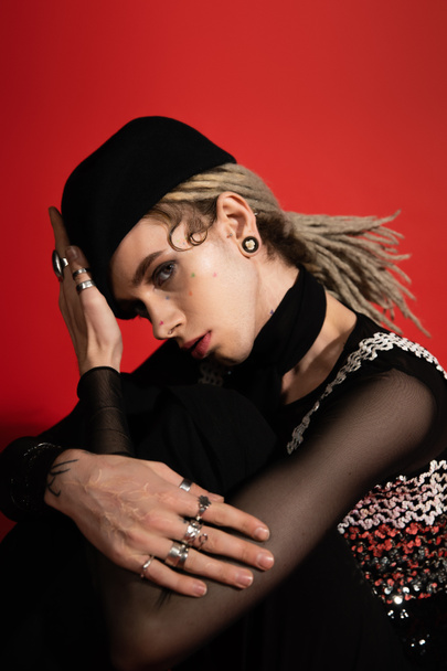 young queer person in silver finger rings and black beret looking at camera isolated on red - Fotoğraf, Görsel