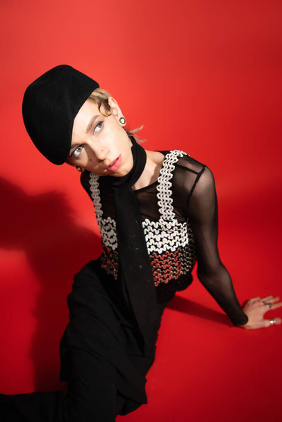 high angle view of trendy nonbinary person in black beret and top with sequins looking away on red background with shadow - Fotoğraf, Görsel