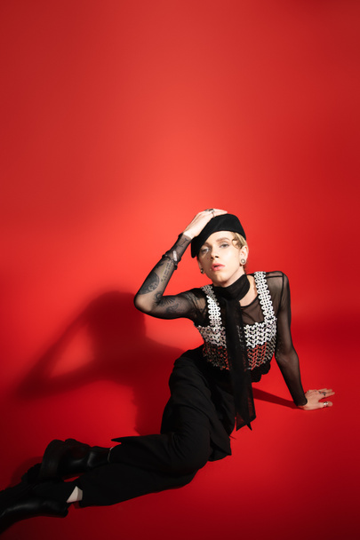 queer person in black and elegant outfit touching beret and looking at camera on red background with shadow - Photo, Image