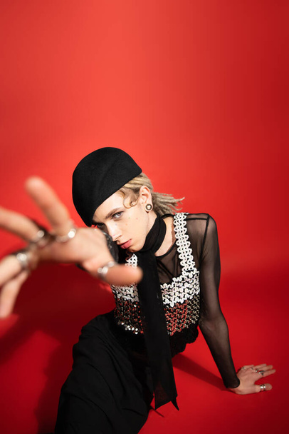 high angle view of stylish queer person in black beret sitting with blurred outstretched hand on red background - Fotó, kép