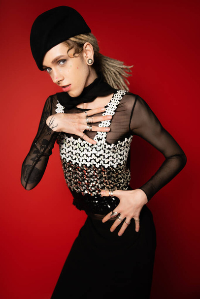 elegant nonbinary model in black beret holding hand on chest while looking at camera on red background - Zdjęcie, obraz