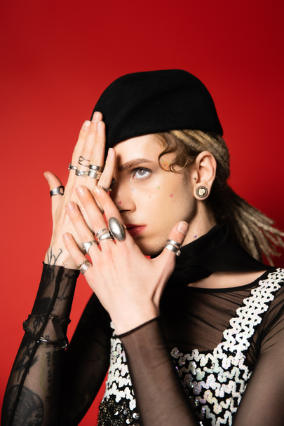 fashionable queer person in black beret and silver rings obscuring face with hands isolated on red - Foto, Imagen