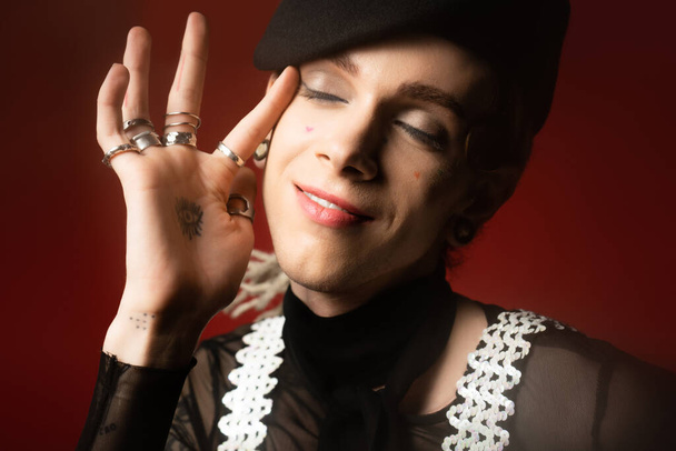 portrait of nonbinary person in stylish attire and silver rings touching face while smiling with closed eyes isolated on dark red - Foto, Imagem