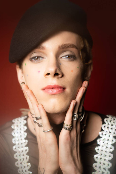 portrait of nonbinary person in black beret and silver finger rings holding hands near face and looking at camera on dark red background - Foto, Imagen