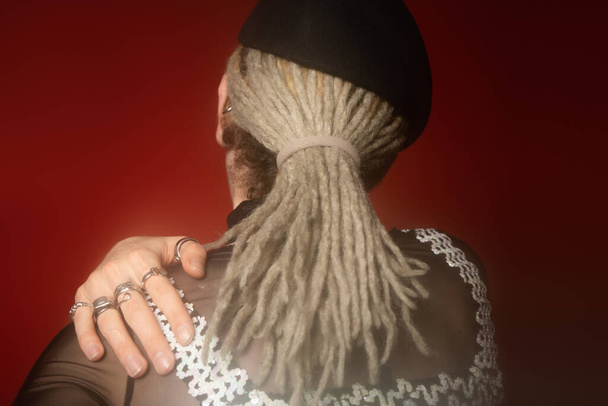 back view of queer person with dreadlocks and silver rings holding hand on shoulder on dark red background - Foto, afbeelding