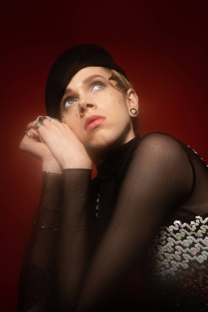 portrait of young queer person in elegant top and black beret looking away on dark red background - Fotó, kép