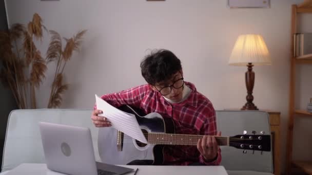 Молодой человек играет на акустической гитаре и смотрит онлайн уроки на ноутбуке, практикуясь дома. Онлайн-обучение, онлайн-классы. Копирование пространства - Кадры, видео