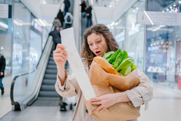 Egy sokkos állapotban lévő nő belenéz egy csekkbe egy bevásárlóközpontban lévő szupermarketből egy mozgólépcső hátterével szemben, és friss termékekkel teli csomagot tart magánál, áremelkedéssel. - Fotó, kép