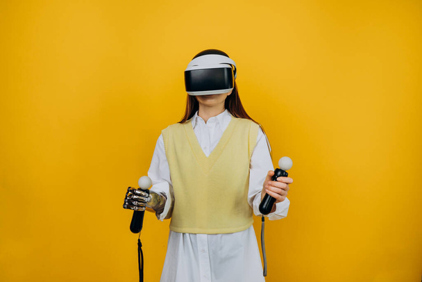 Ξανθιά κυρία σε λευκό πουκάμισο με βιονικό βραχίονα χρησιμοποιώντας γυαλιά εικονικής πραγματικότητας και joysticks παίζει παιχνίδι σε κίτρινο φόντο closeup - Φωτογραφία, εικόνα