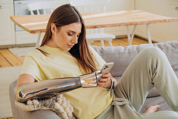 Happy woman with a bionic prosthetic arm sits on the sofa with a phone and communicates online with friends or surfs Internet or makes online purchases in a yellow T-shirt against the background of a - Photo, Image