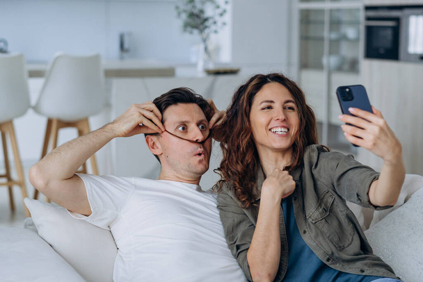 Χαρούμενο ερωτευμένο ζευγάρι που βγάζει μια selfie στο τηλέφωνο ενώ κάθεται στον καναπέ μαζί, βγάζοντας αστεία μουστάκια από τα μαλλιά τους και βγάζοντας φωτογραφίες - Φωτογραφία, εικόνα