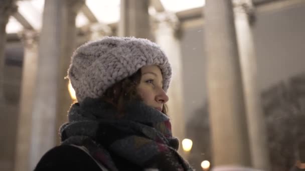 Eşarba sarılmış örülü şapkalı bir kadın akşamleyin fenerlerin arka planında, yan görüş ile caddede yürür. Genç ve güzel bir kız bir kış akşamı ofisten çıkıp yürür.. - Video, Çekim
