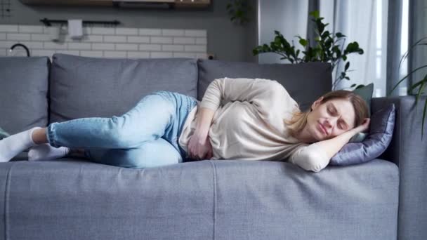 Sick young blond woman suffering from stomach ache laying on the couch holding belly feeling gastritis or menstrual pain in the living room at home Gynecology problem Painful periods days concept  - Záběry, video