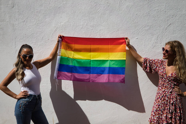 サングラスをかけた2人の若い女性がゲイのプライド旗を手に持っています。女性たちはゲイのプライドの日を喜んでいる。平等概念同性愛lgtb - 写真・画像