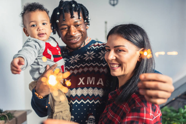 Smiling multicultural parents and their toddler with Christmas sparkling fire sticks. High quality photo - Zdjęcie, obraz