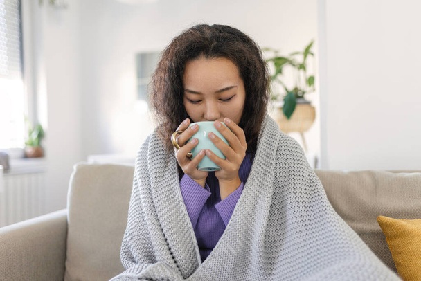 Sick young Asian woman sitting at home in bed with hot cup of tea and handkerchief. Seasonal colds, cough, runny nose, viral infections, home treatment - Foto, imagen