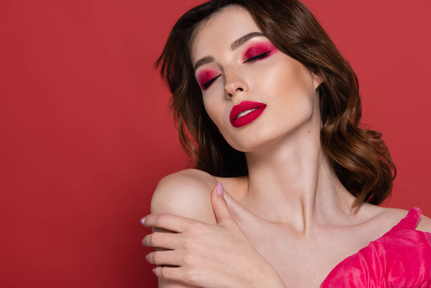 portrait of young woman with magenta color eye shadow and glove embracing herself isolated on pink  - Photo, Image