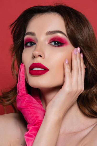 portrait of young woman with magenta color glove looking at camera while touching face isolated on pink  - Photo, Image