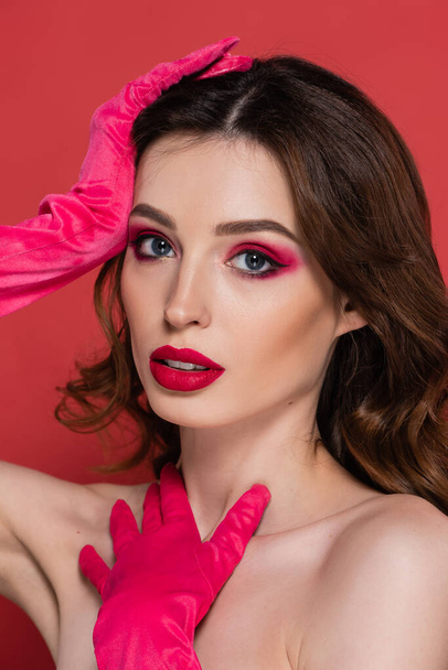portrait of brunette woman with magenta color eye makeup looking at camera while posing in gloves isolated on pink  - Foto, immagini