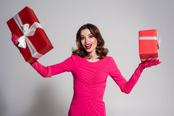 amazed young woman in magenta color dress and gloves holding presents on grey  - Foto, immagini