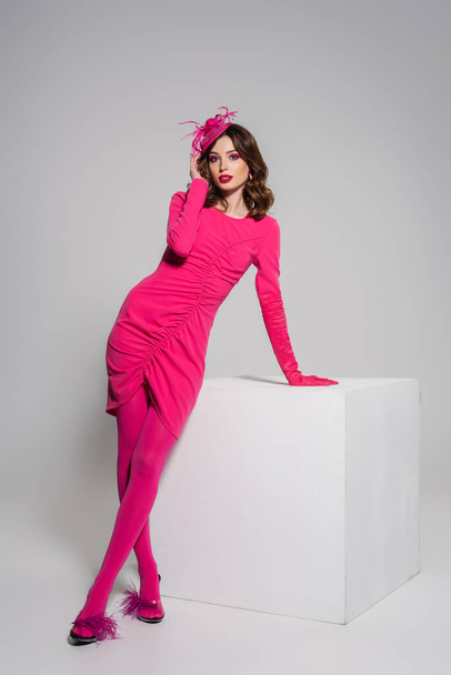 full length of young woman in magenta color dress and elegant hat with feather posing near white cube on grey  - Foto, Imagem