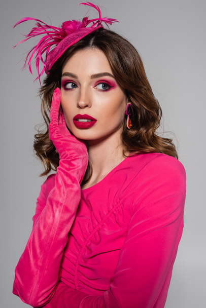 dreamy young woman in magenta color dress and elegant hat with feather looking away isolated on grey  - Fotografie, Obrázek