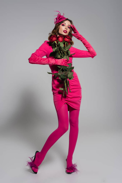 full length of young woman in magenta color gloves and hat with feather holding red roses on grey  - Fotoğraf, Görsel