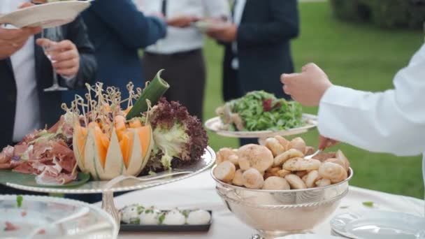 Servant in uniform serving guest from organised banquet table putting food delicacy assortment on plate, bees and insects ruining smelling food outdoor, problem of unhygienic food on open-air catering - Záběry, video