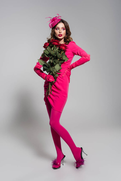 full length of young woman in magenta color gloves and hat posing with hand on hip while holding red roses on grey  - Fotoğraf, Görsel