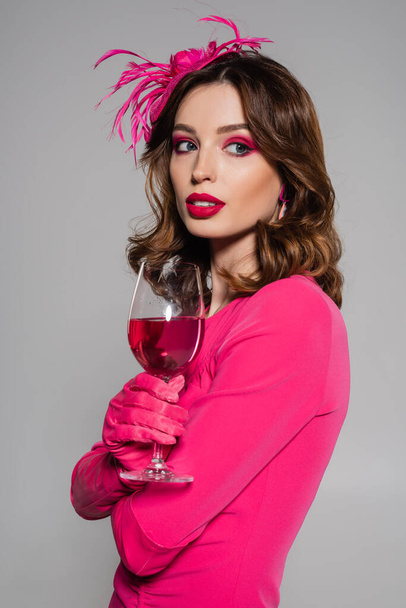 portrait of brunette woman in elegant hat and magenta color dress holding glass with alcohol drink isolated on grey  - Foto, afbeelding