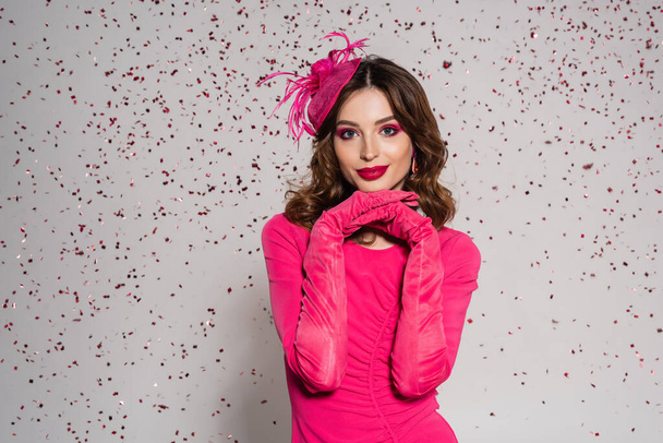 cheerful woman in elegant hat and magenta color dress posing on grey background with falling confetti  - Fotografie, Obrázek