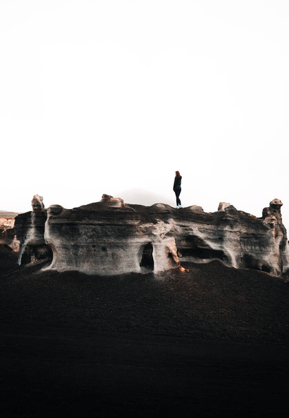 Minimalistic vertical photo of woman tourist standing on the top of rock in desert. Stratified rocks (teseguite - Lanzarote) with traveler on top in dramatic and moody atmosphere.  - Fotoğraf, Görsel